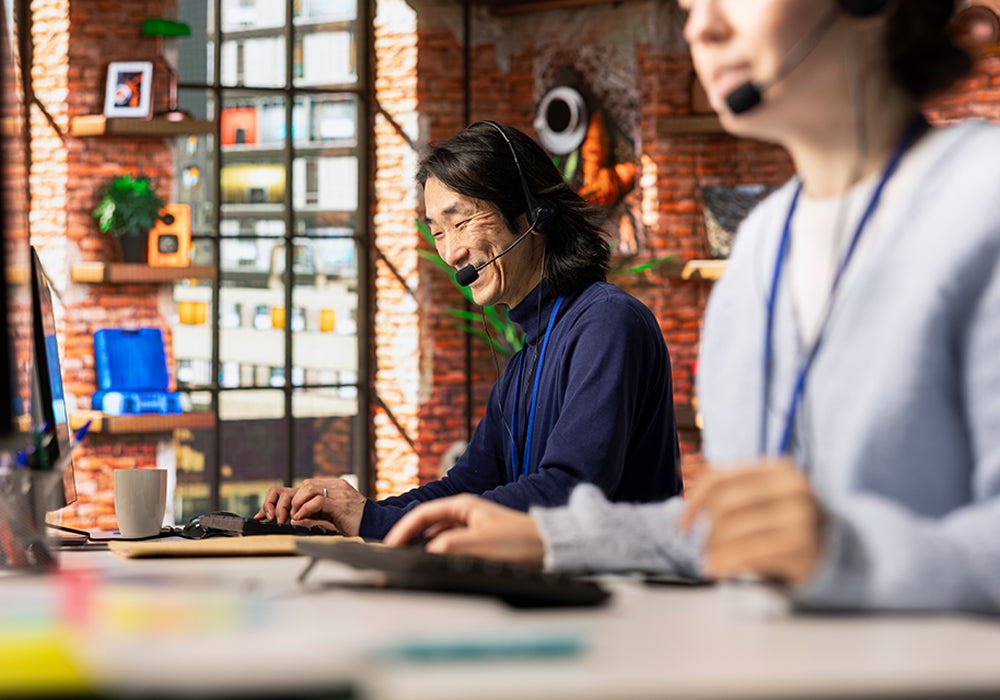 Two people working at a desk with computers and headsets in an office setting.
